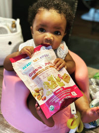Child sitting in pink baby chair holding a bag of RSF! Organic Puffs